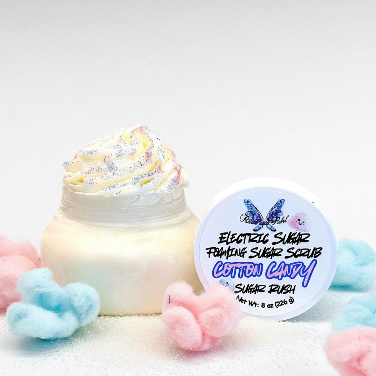 Electric Sugar Foaming Sugar Scrub by Ritual and Rebel clear plastic jar with cream colored whipped body butter with white round lid and pink and blue cotton candy and sugar around base of jar 8 oz