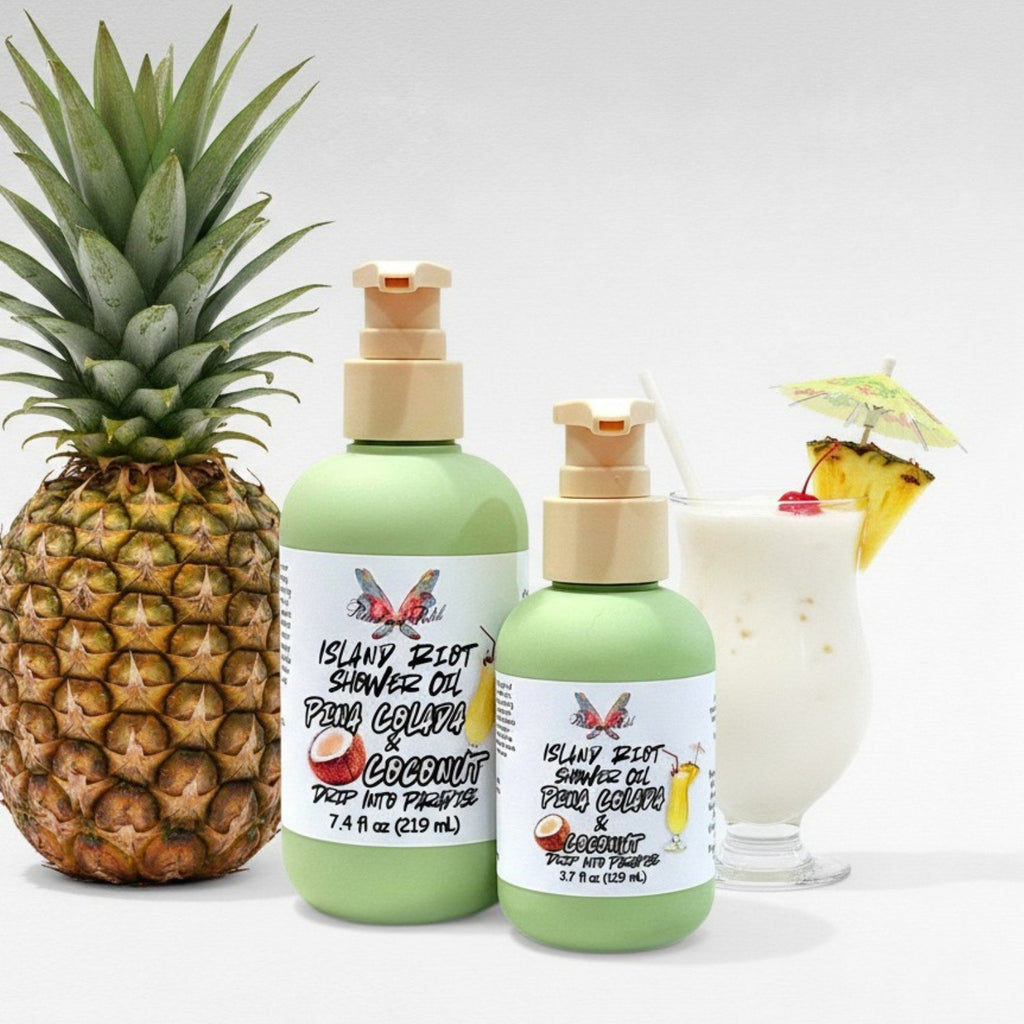 Island Riot Shower Oil by Ritual and Rebel with two green bottles 7.4 & 3.7 fl oz with a pineapple and a pina colada drink in the background.